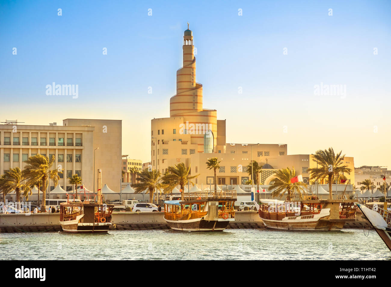 Traditional wooden dhow anchored at Dhow Harbor in Doha Bay with spiral mosque and minaret in the background at sunset. View from Corniche promenade Stock Photo