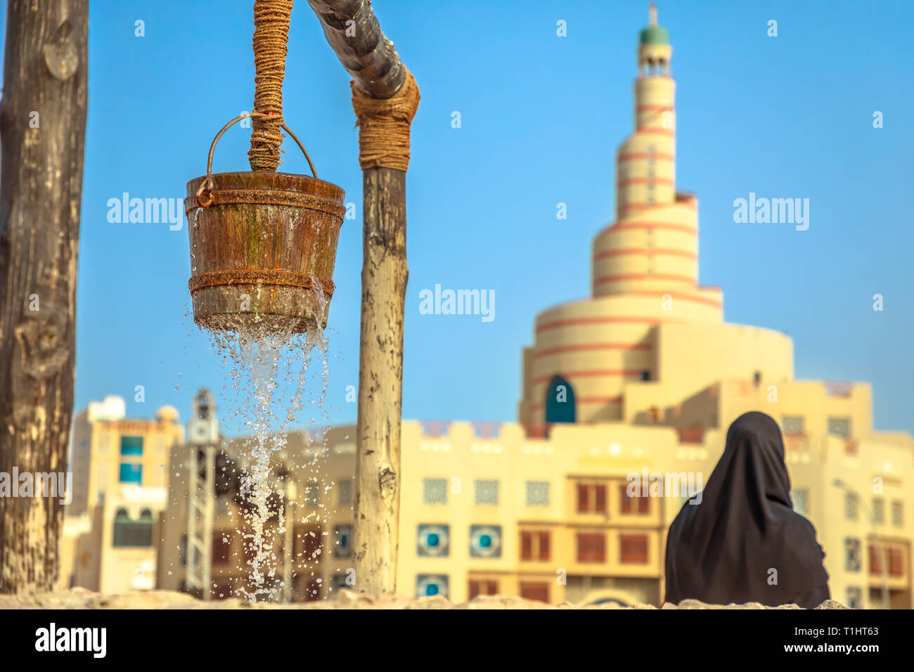 Closeup of flowing water at old fountain at Souq Waqif in Doha center with woman behind with dressed abaya and Doha Mosque with minaret on blurred Stock Photo