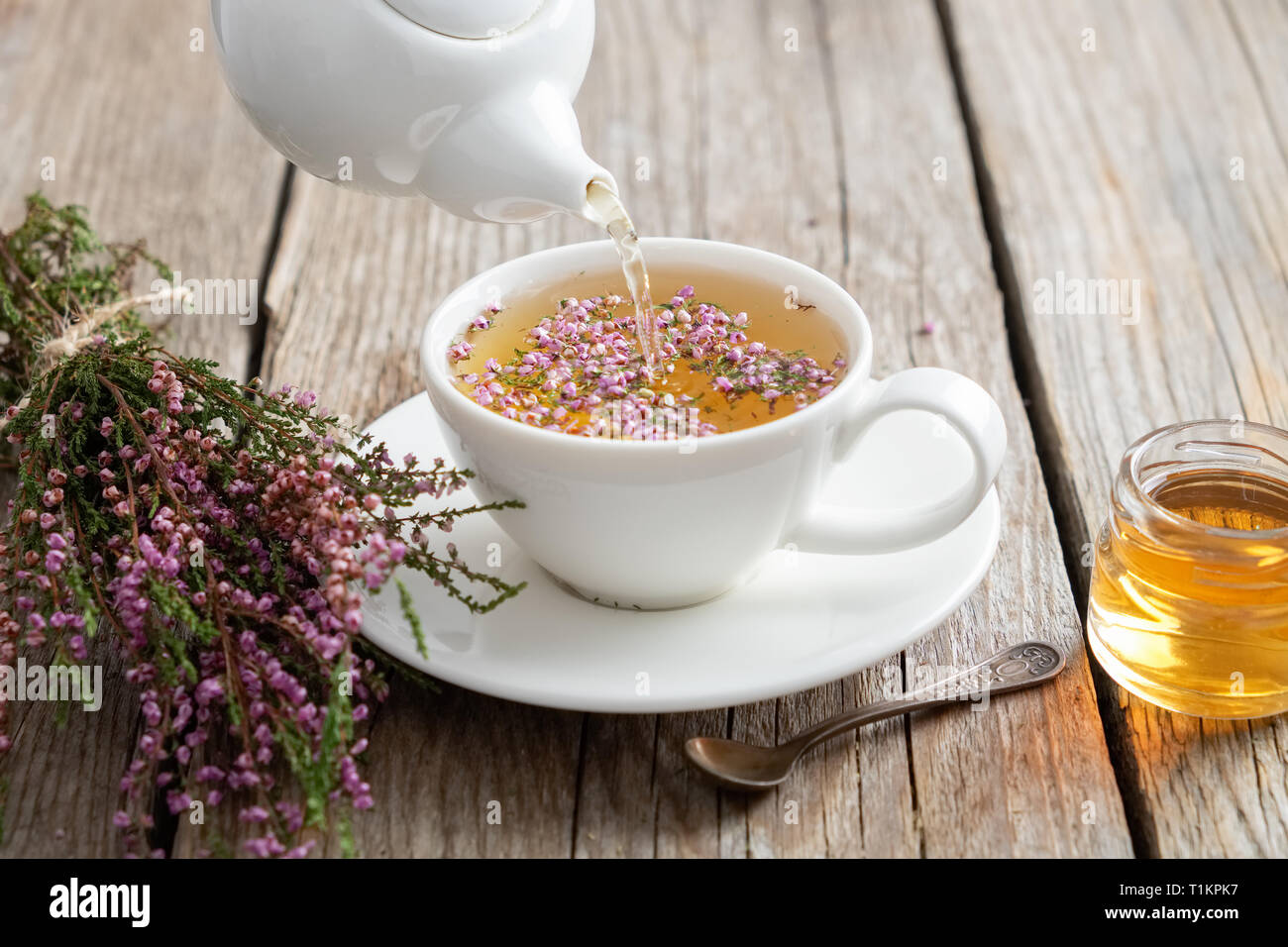 Healthy heather tea poured into white cup. Teapot, small honey jar and heather bunch. Stock Photo