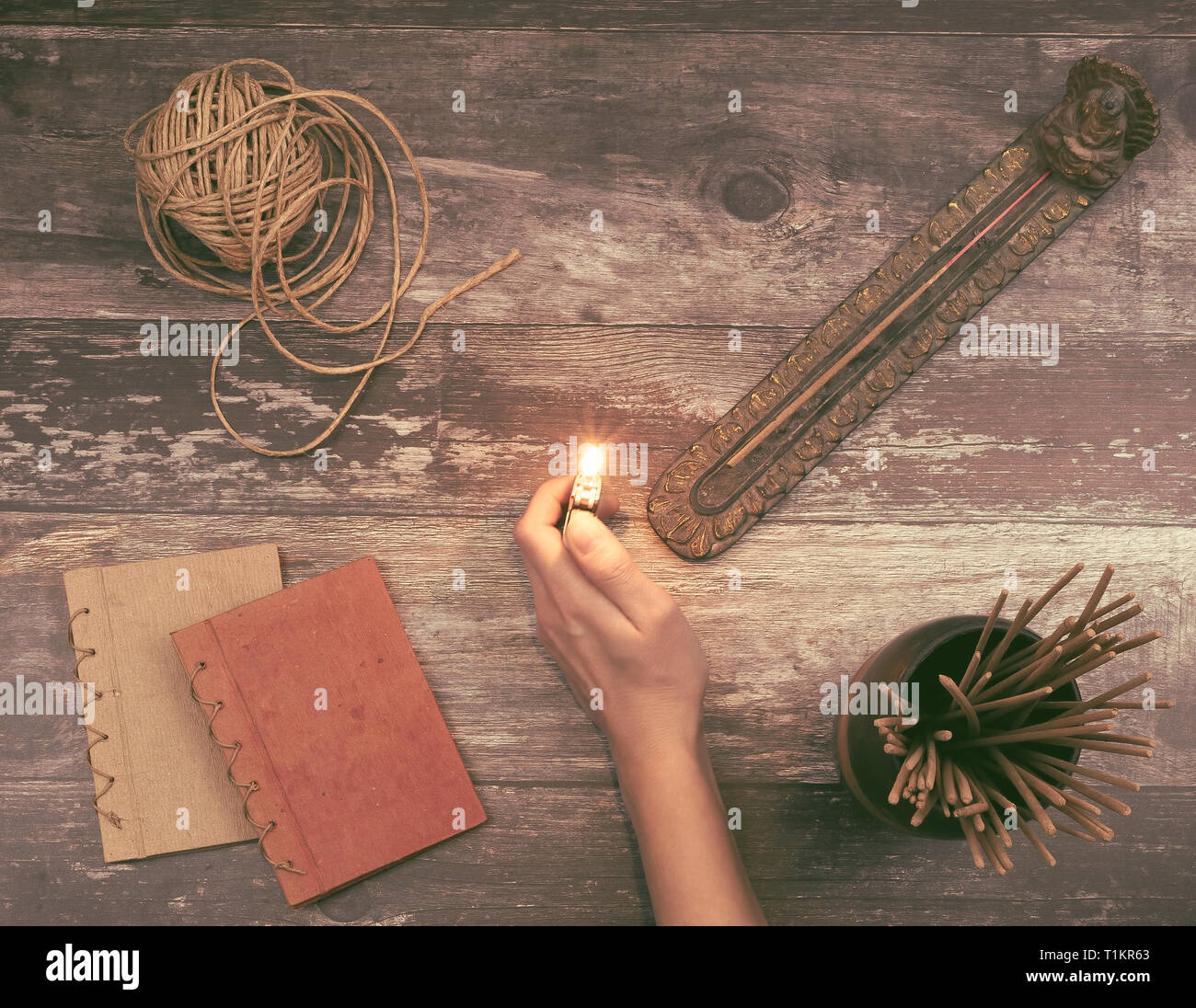 Woman hand lights an incense stick from a Buddha holder on a vintage natural wooden surface with books, hemp twine and many aromatic incense sticks Stock Photo