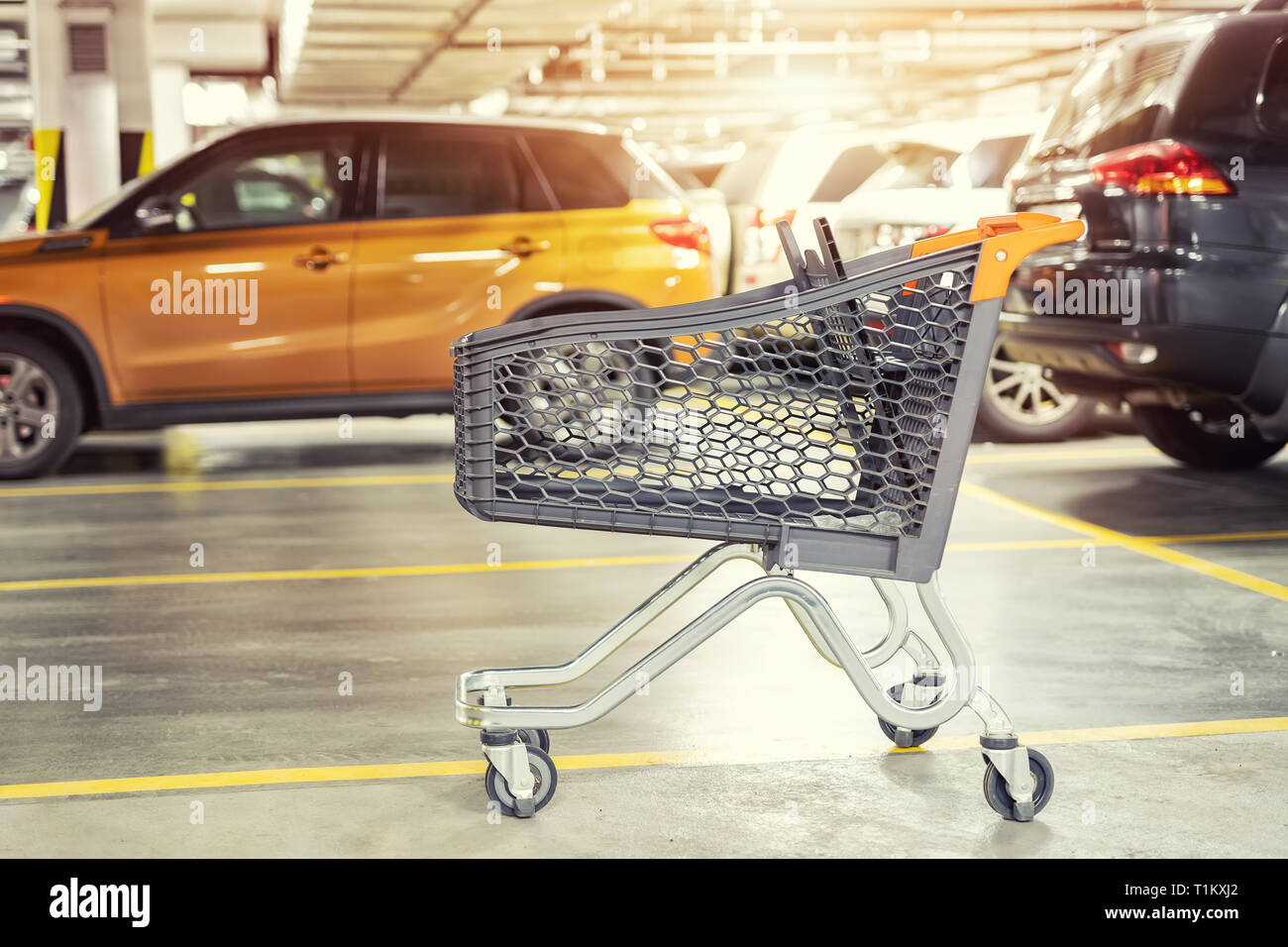 Modern plastic empty shopping cart left at roofed underground parking of shopping mall. Supermarket trolley with blurred parked cars on background. Co Stock Photo