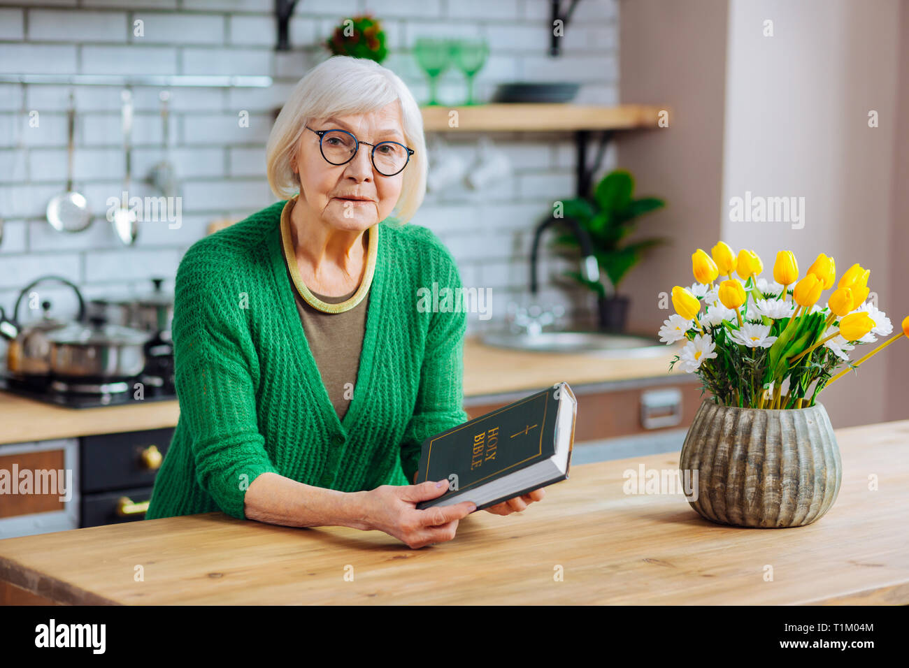 Beautiful grey-haired woman holding a Holy Bible book at kitchen Stock Photo