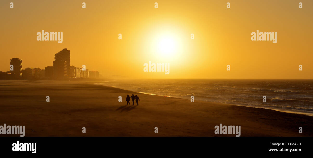 Panorama of a sunset along the North Sea beach of Ostend with the silhouette of three people walking during a sand storm, West Flanders, Belgium. Stock Photo