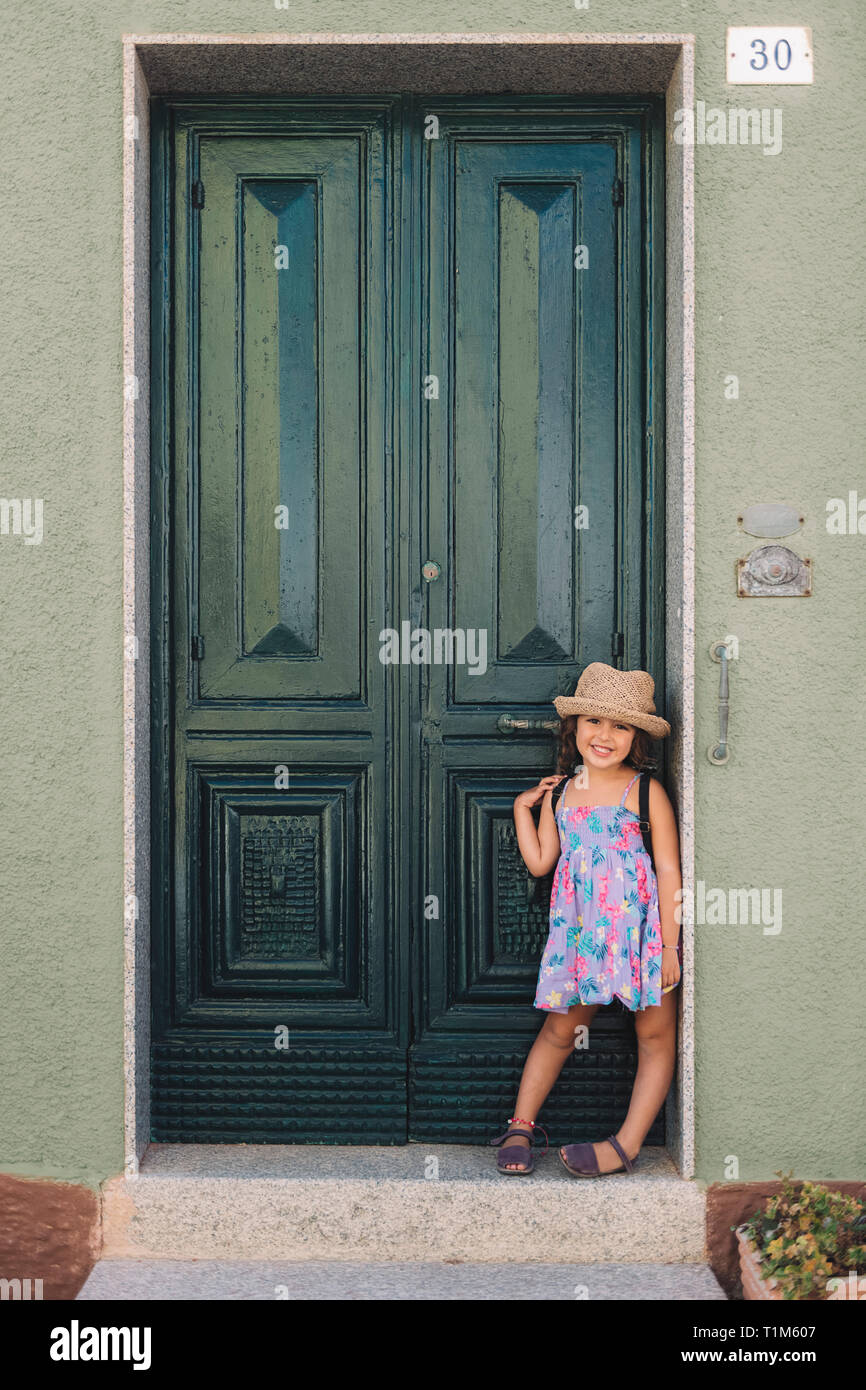 Little girl poses smiling leaning against the green door of her house ...