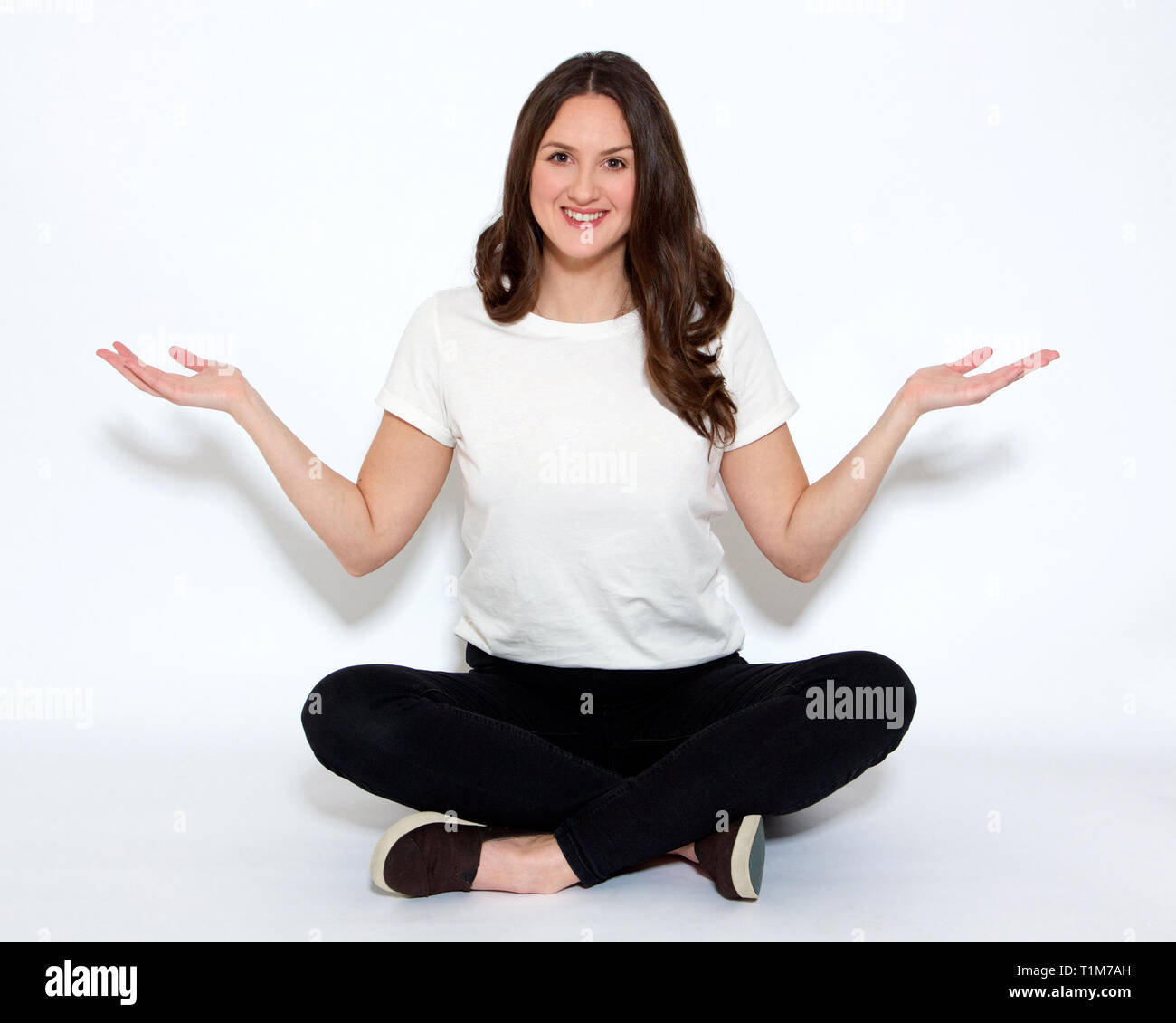 Woman sitting cross-legged holding her hands out sideways wearing a white top and black jeans Stock Photo