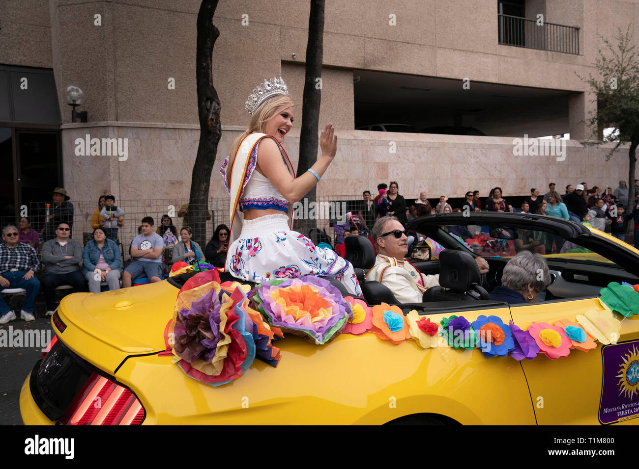 Young woman wearing crown sits on back of convertible car and waves to
