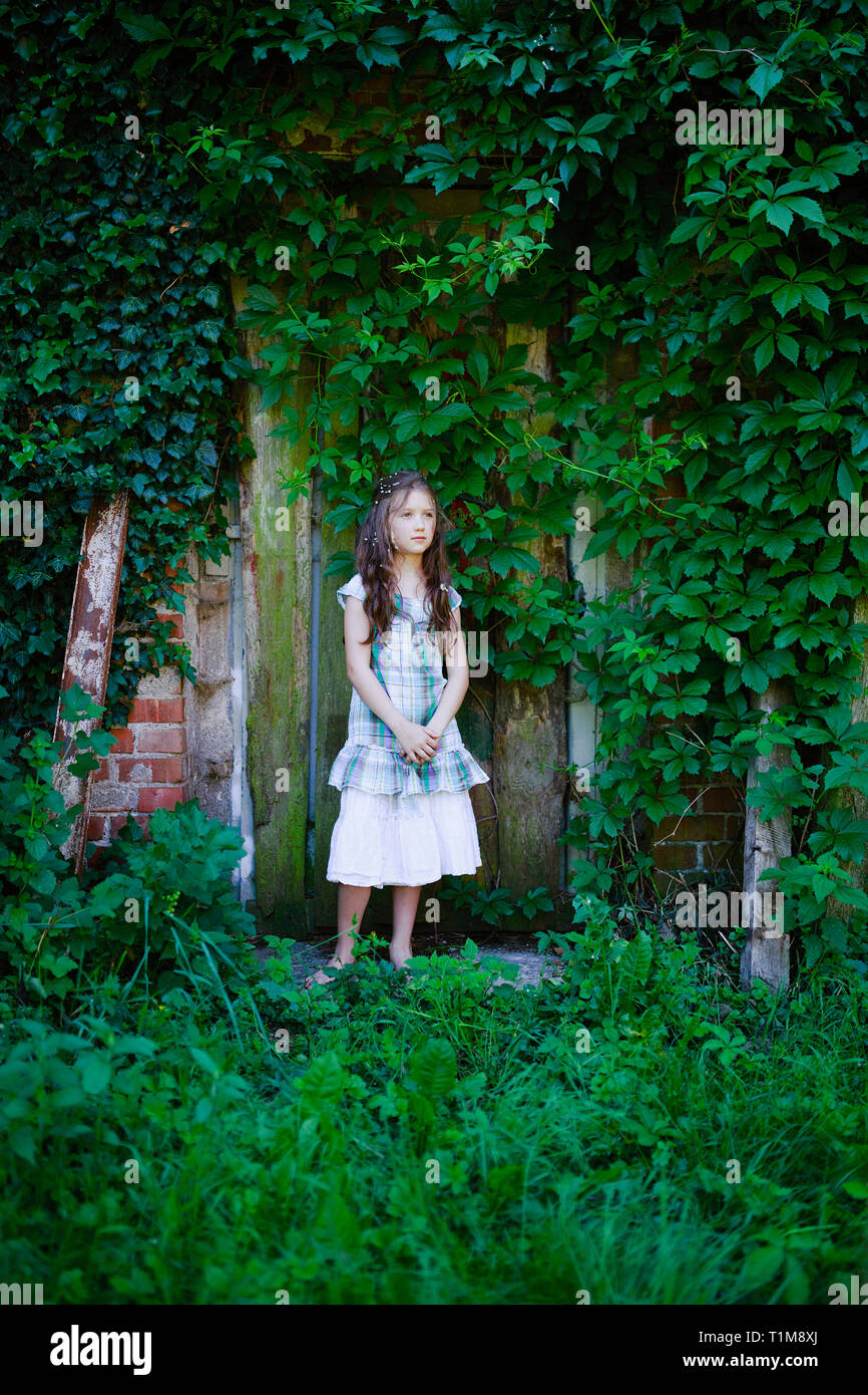 Serene girl in dress standing at rustic door covered with green ivy Stock Photo