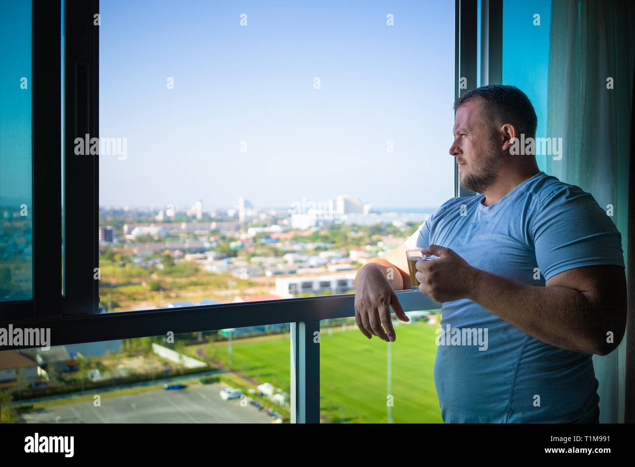 male drinks coffee and looks through panoramic window smiles in the morning Stock Photo