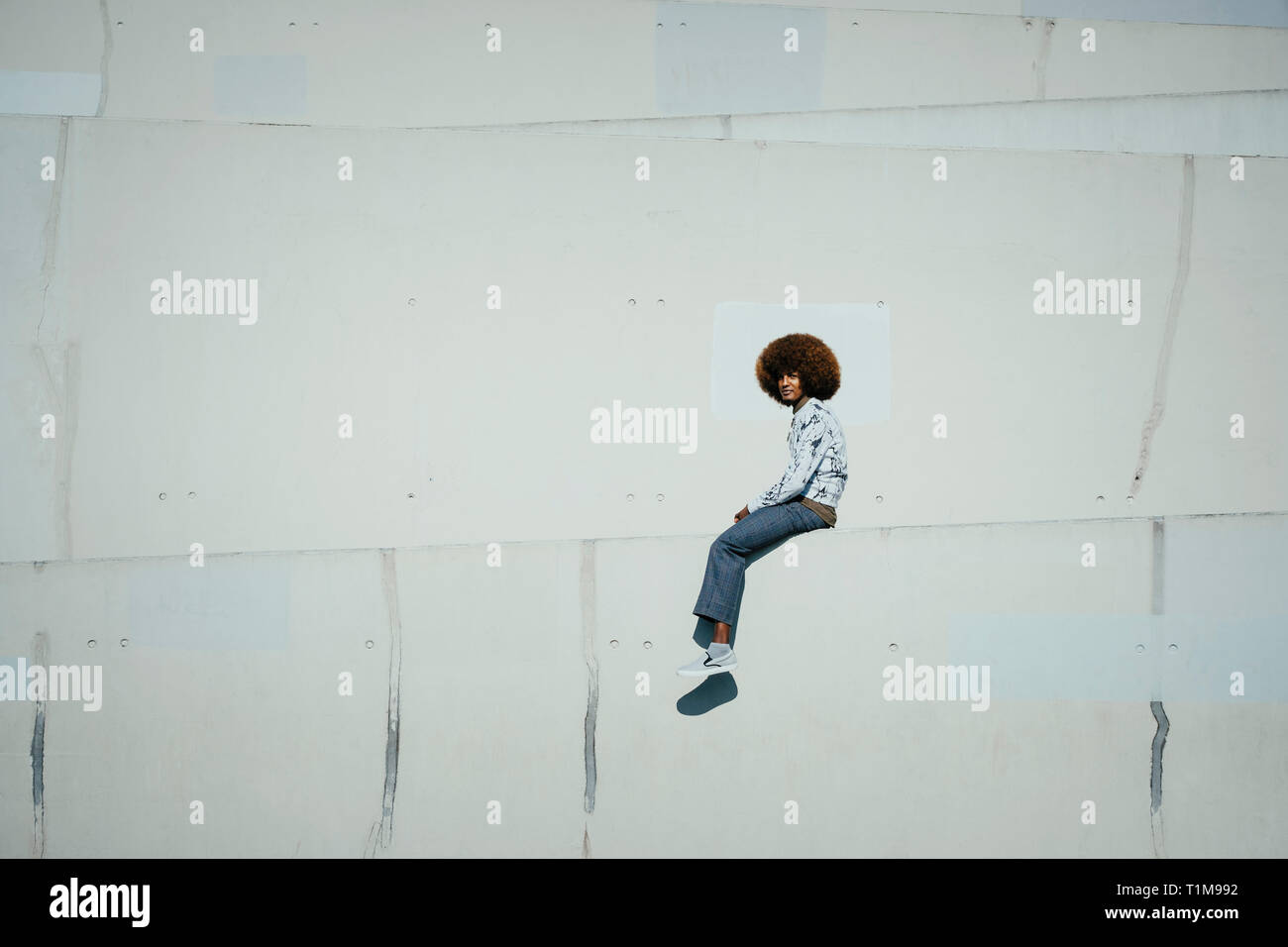 Portrait confident young man with afro sitting on sunny urban wall Stock Photo