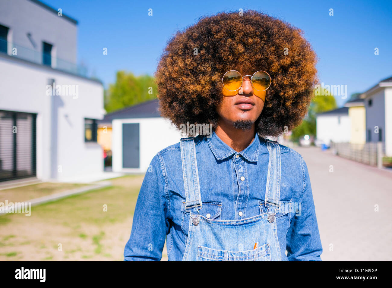 Portrait cool young man with afro in denim overalls standing on sunny road Stock Photo