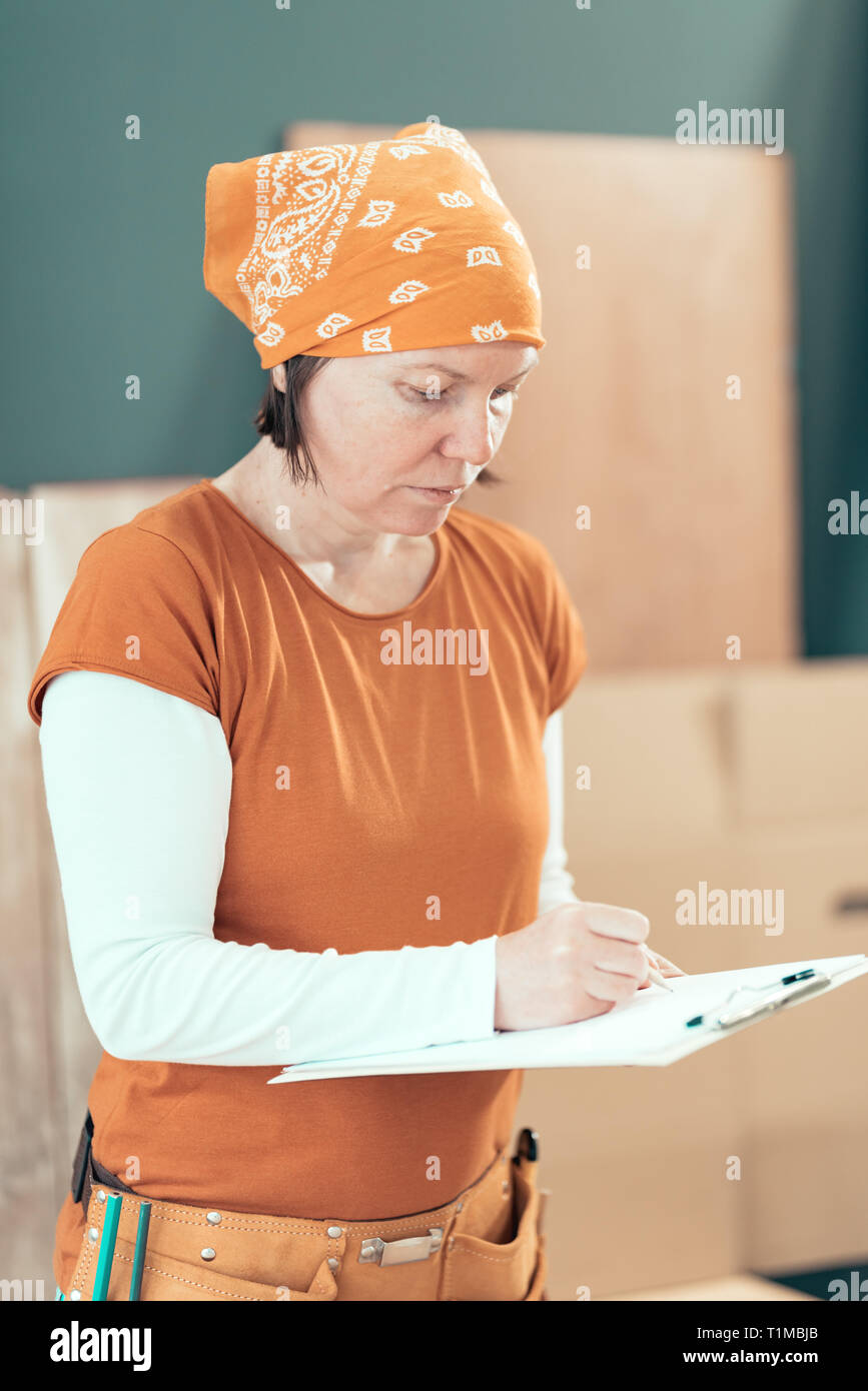 Female carpenter wearing bandanna is writing project notes on clipboard note paper in her small business woodwork workshop Stock Photo
