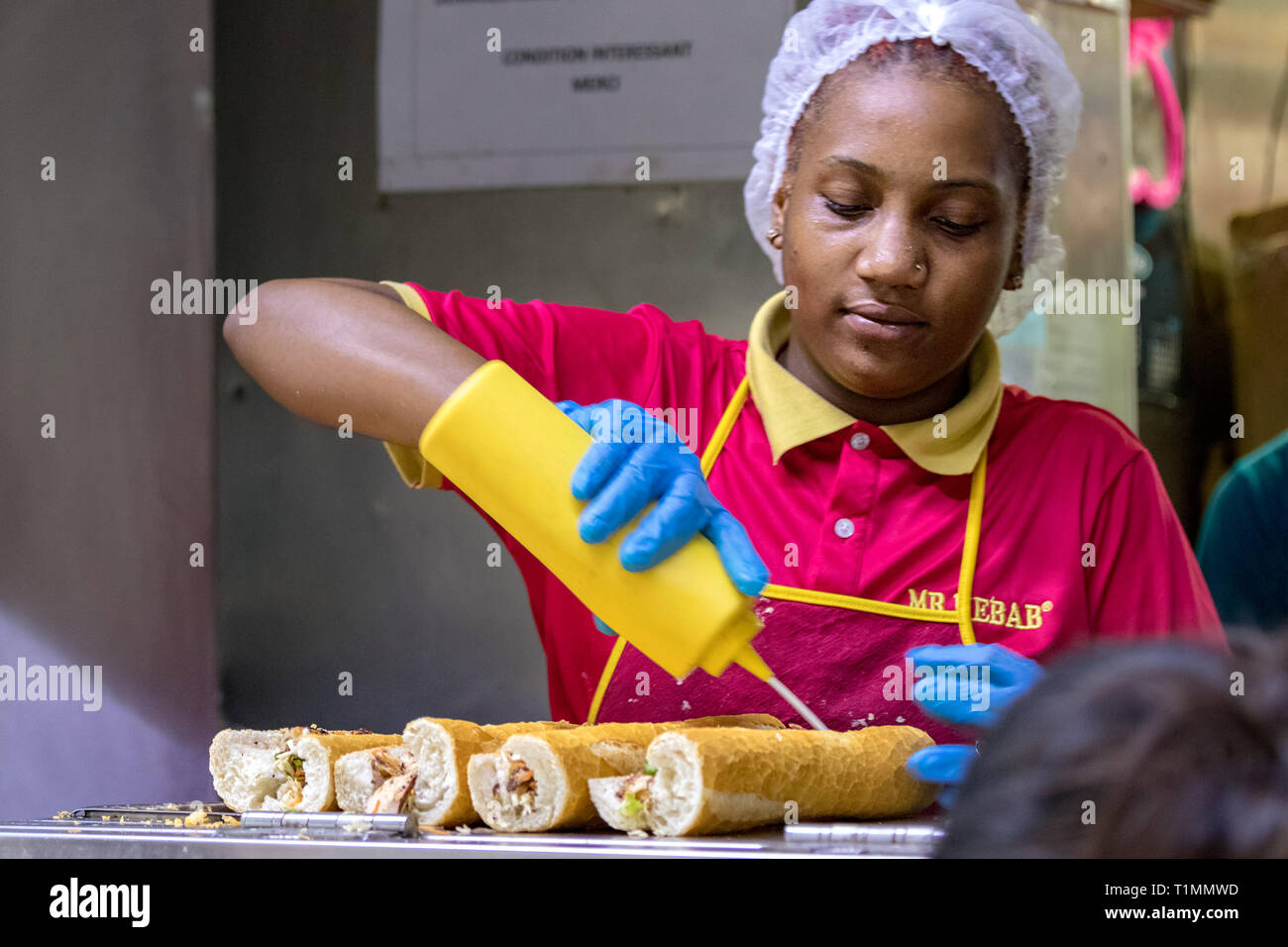 Port Louis, Mauritius - January 30, 2019: A woman working in a fast food restaurant filling with sauce a row of sandwiches in the Bus Terminal next to Stock Photo