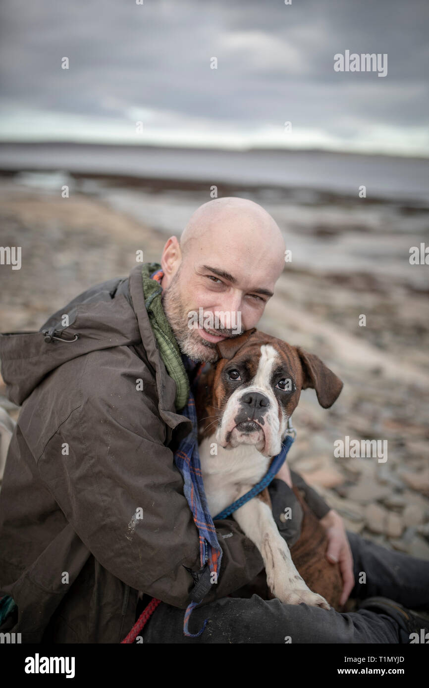 A front-view shot of a mature caucasian man sitting on a beach with his brown and white boxer dog, they are embracing eachother on the cold day in the Stock Photo