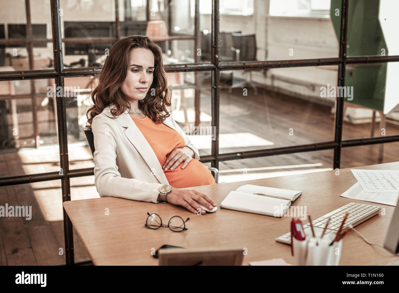 Focused long-haired woman keeping her hand on pregnant belly Stock Photo