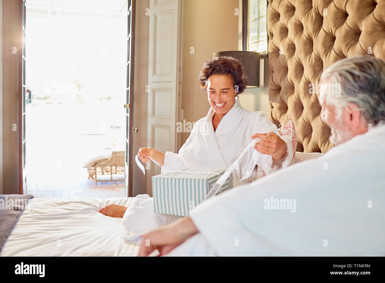 Husband watching happy wife opening gift on hotel bed Stock Photo