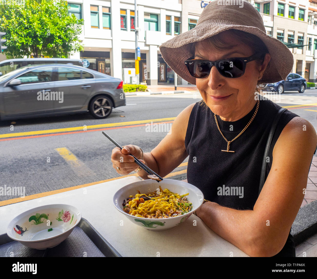 Caucasian female tourist eating traditional Singaporean breakfast bowl of noodles with minced pork and mushrooms al fresco North Bridge Rd Singapore. Stock Photo