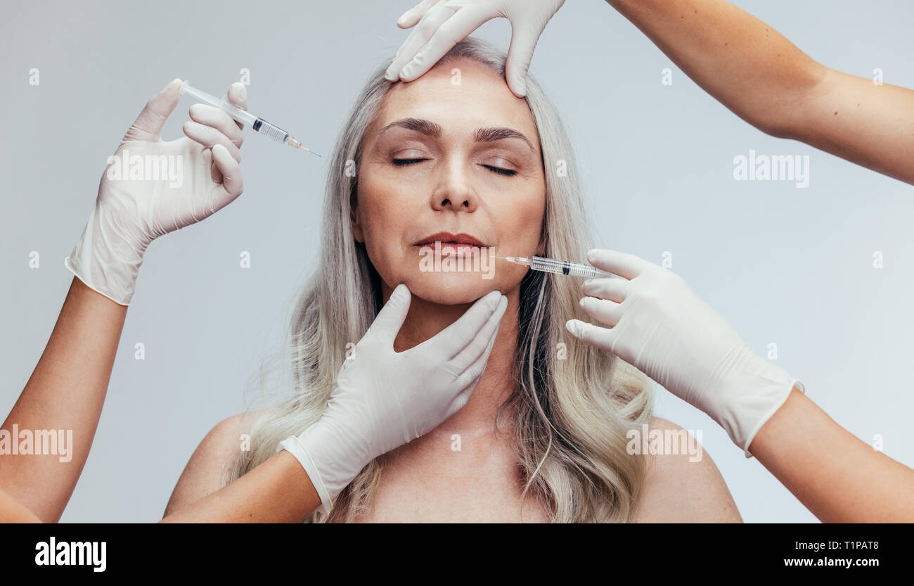 Mature woman getting anti wrinkles aesthetic treatment on face by cosmetic doctors. Senior female getting injection on her face by beauticians over gr Stock Photo
