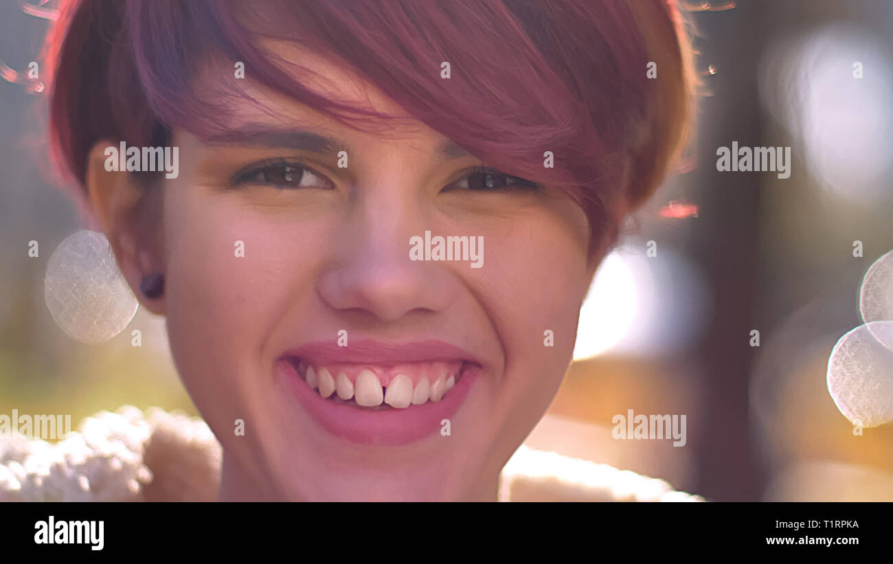 Close-up portrait of young cool caucasian pink-haired girl watching into camera with extreme happiness on sunny park background. Stock Photo