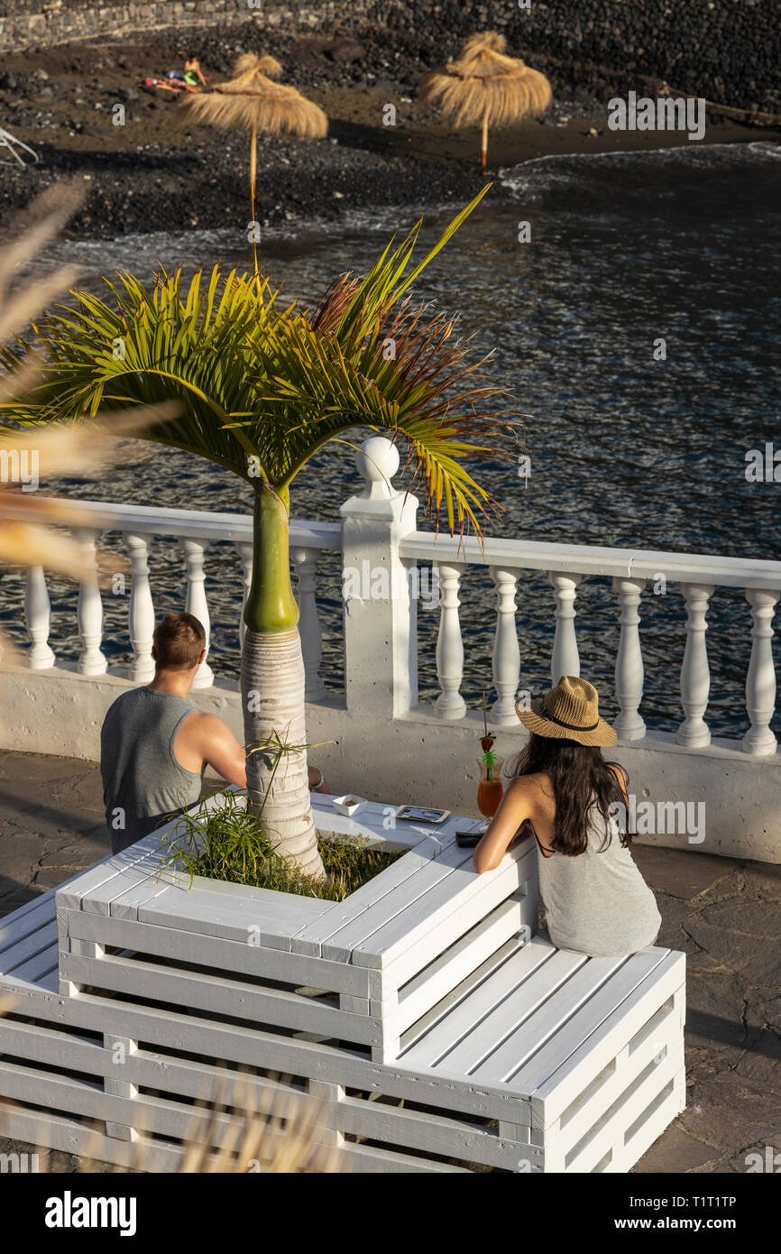 Young couple enjoying a cocktail sitting on a wooden seat by a palm tree on the harbour wall at the Playa Chica, Puerto de Santiago, Tenerife, Canary  Stock Photo