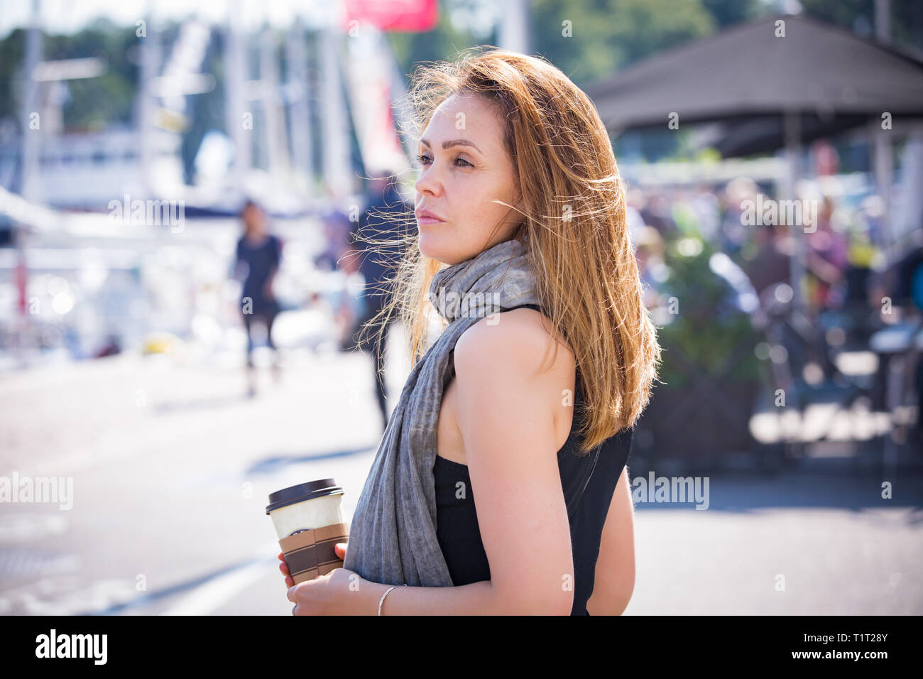 Portrait of smiling stylish middle aged woman walking along sea bay. Sailboats in harbour, seaside. Sunny summer day. Wind in hair, drinking coffee Stock Photo