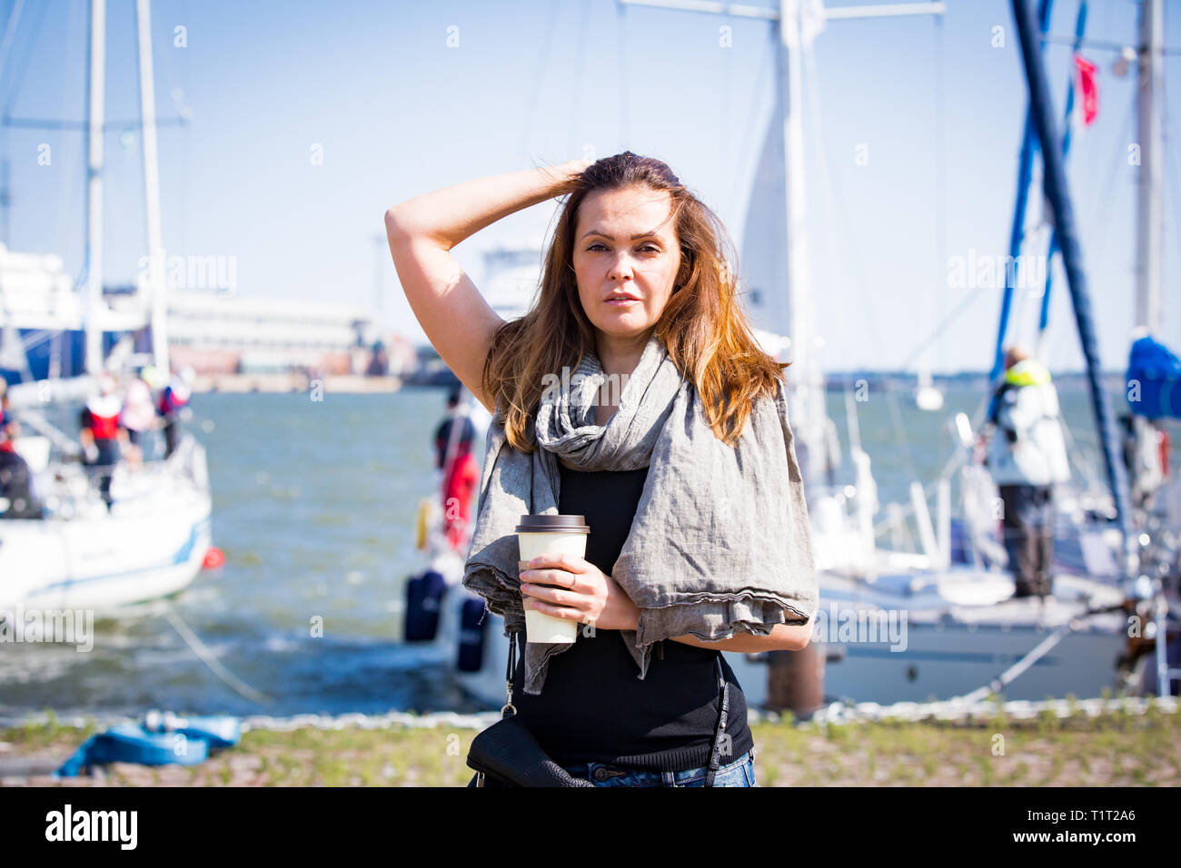Portrait of smiling stylish middle aged woman walking along sea bay. Sailboats in harbour, seaside. Sunny summer day. Wind in hair, drinking coffee Stock Photo