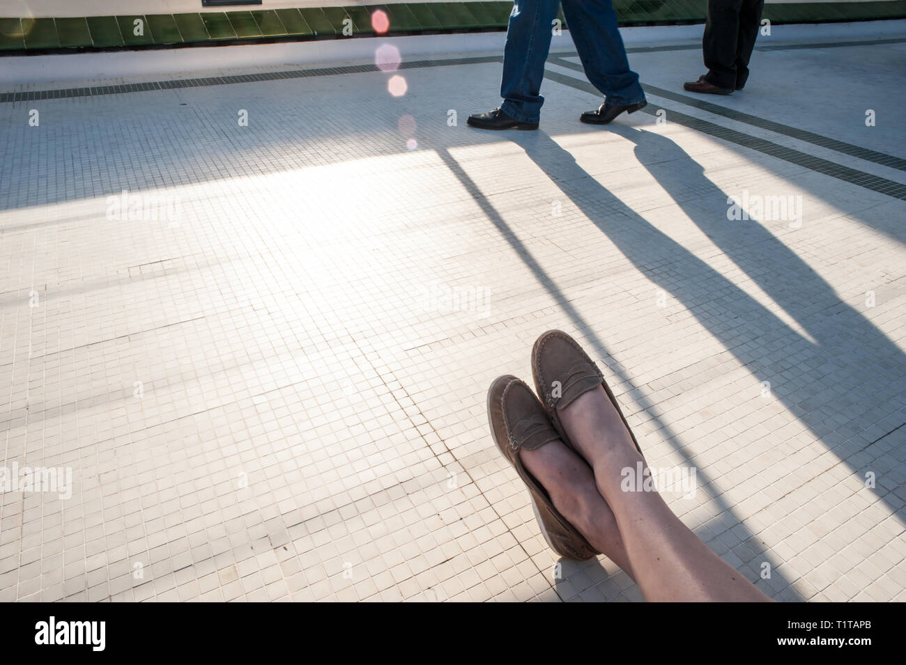 A woman relaxes in the evening sun while passers by cast long shadows. Stock Photo