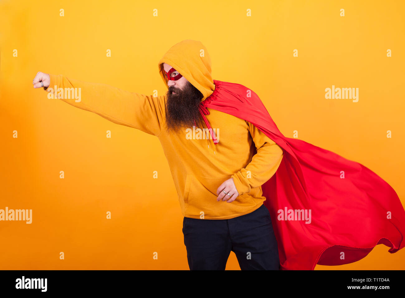 Bearded hipster dressed in superhero costume flying away to save the world over yellow background. Stock Photo