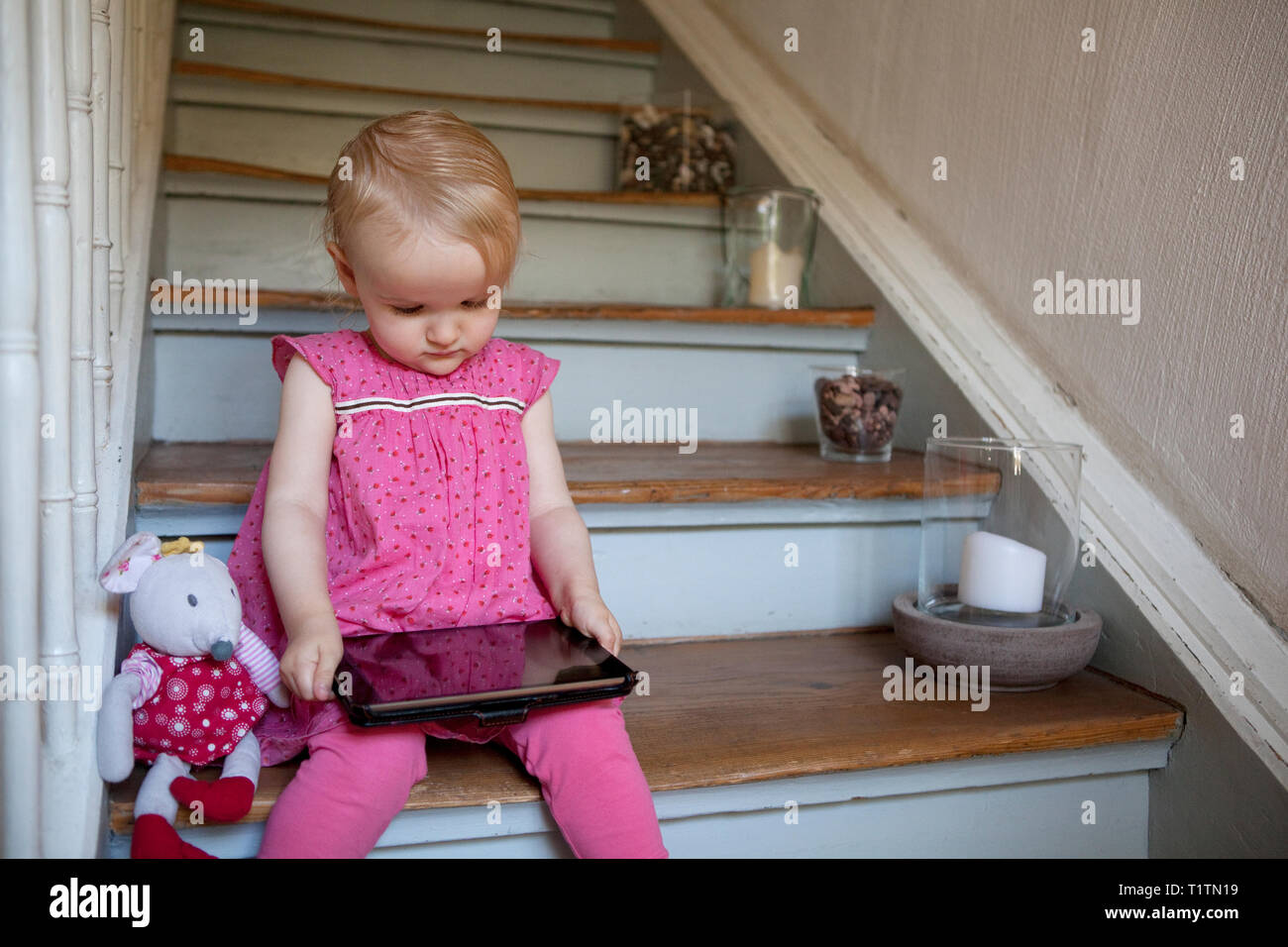 Toddler sitting on steps in a house and looking at a portable tablet device Stock Photo