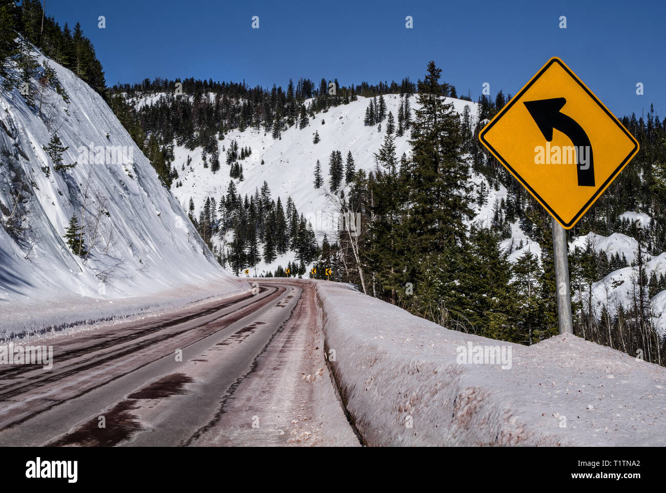 Left Curve Sign:  Motorists are warned of a sharp turn ahead on a snowy mountain road in Montana. Stock Photo