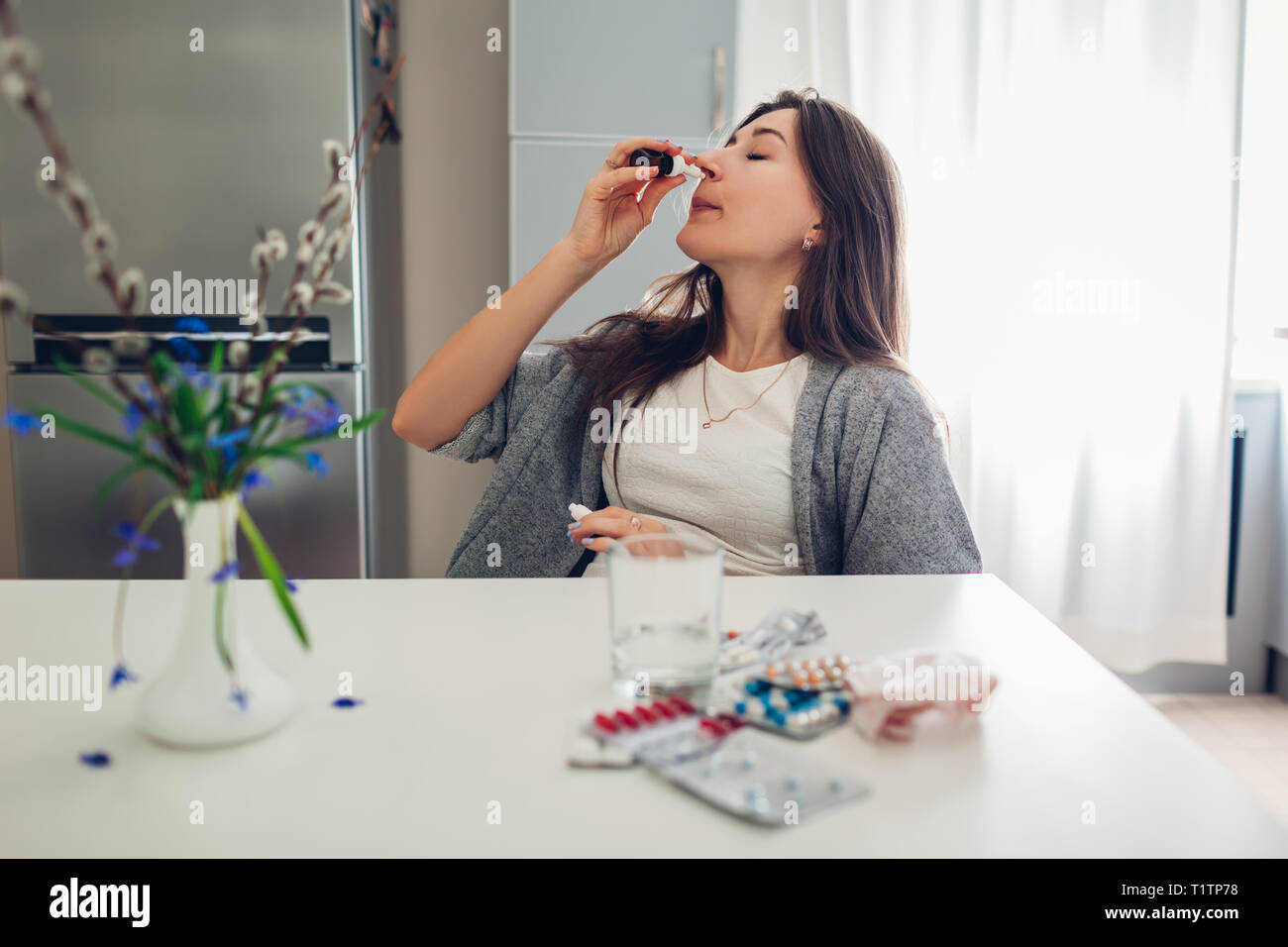 Spring allergy. Tired woman using nasal drops against seasonal allergy and taking pills on kitchen at home. Healthcare and medicine Stock Photo