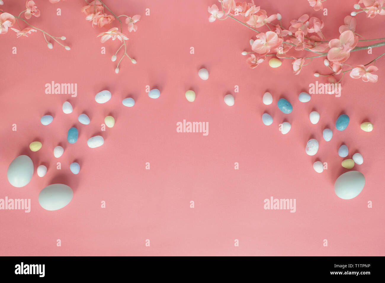 Pastel Easter eggs, malt candy covered chocolate eggs, and flower blossoms over a coral colored background with copy space. Image shot from top view. Stock Photo