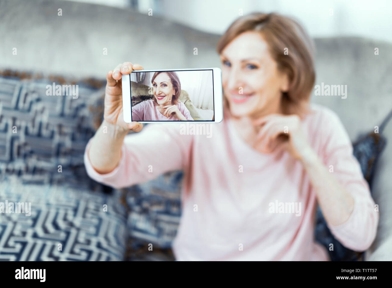 Elderly Woman With a Phone in Her Hands in Good Mood is Resting at Home in the Living Room. Stock Photo