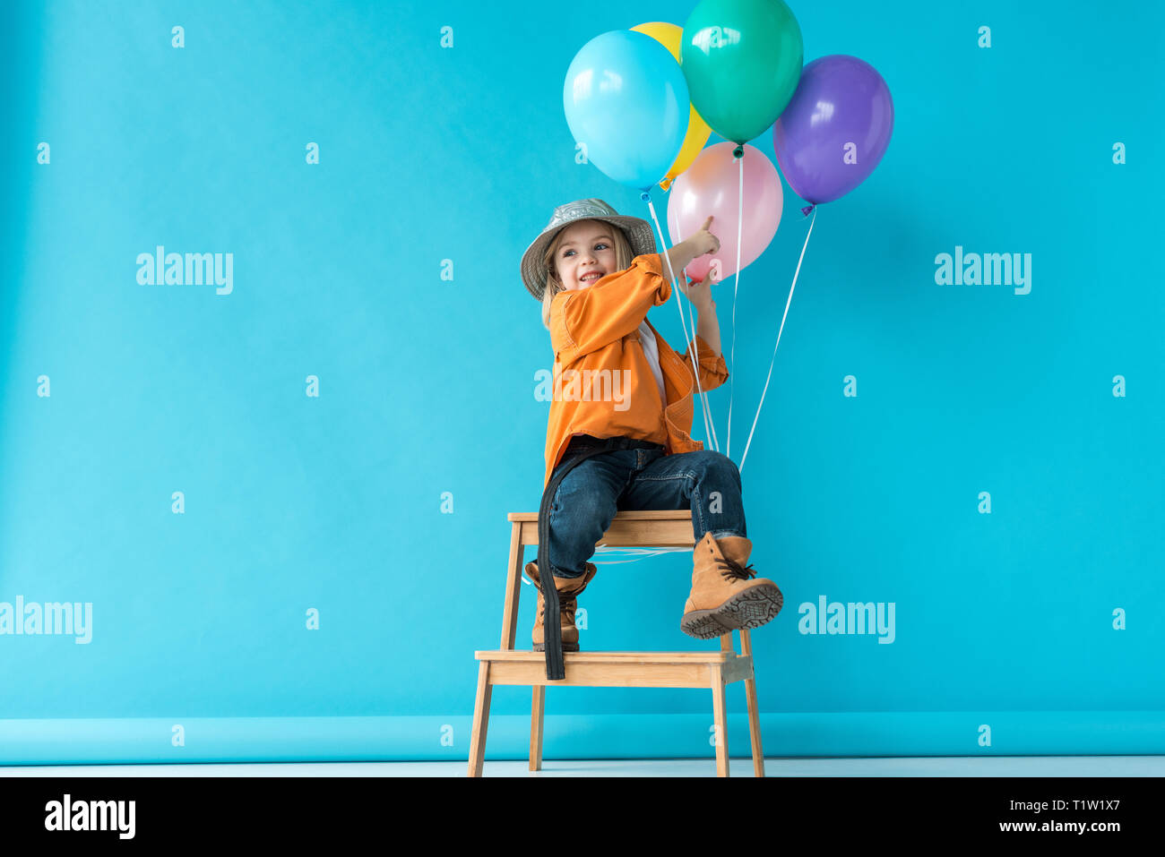 cute kid in jeans and orange shirt sitting on stairs and pointing with finger at balloons Stock Photo