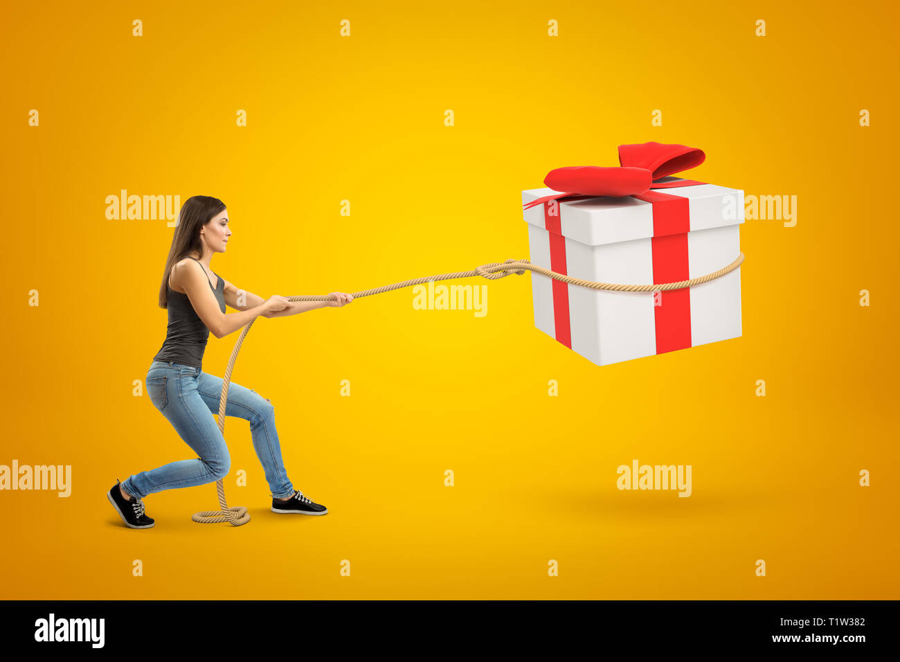 Side view of young woman standing with bent knees and pulling big gift box in air which she has lassoed, on yellow background. Stock Photo