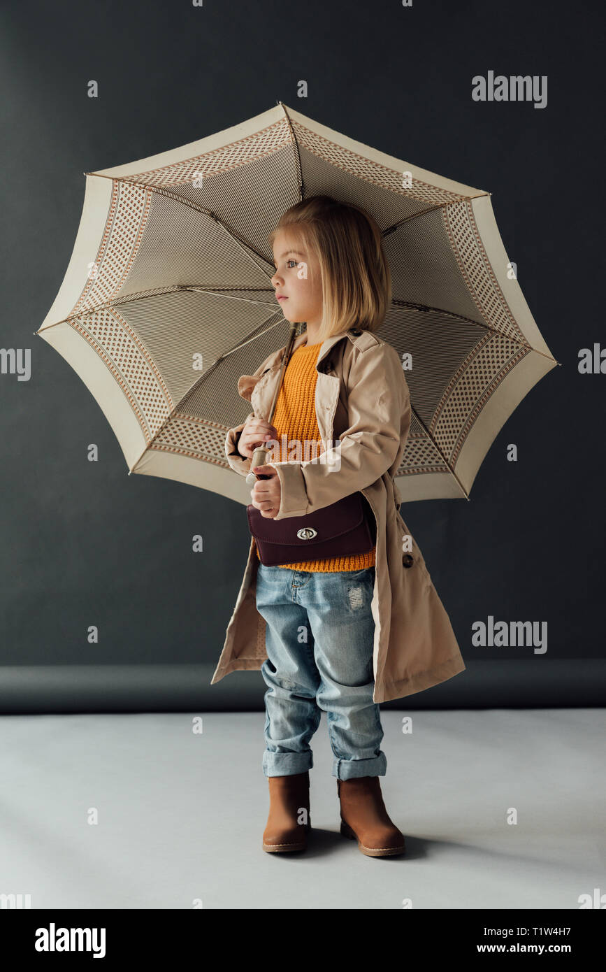 serious child in trench coat and jeans holding umbrella and looking away Stock Photo