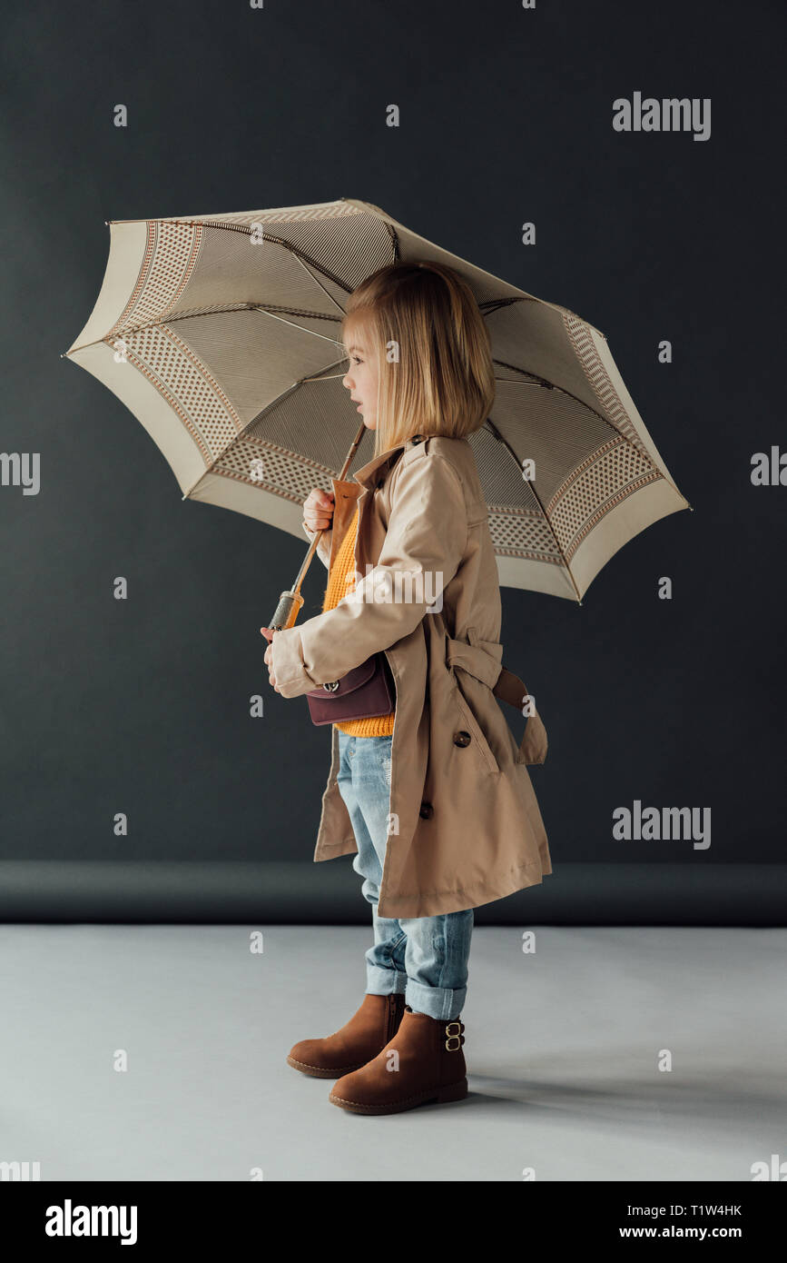 side view of child in trench coat and jeans holding umbrella on black background Stock Photo