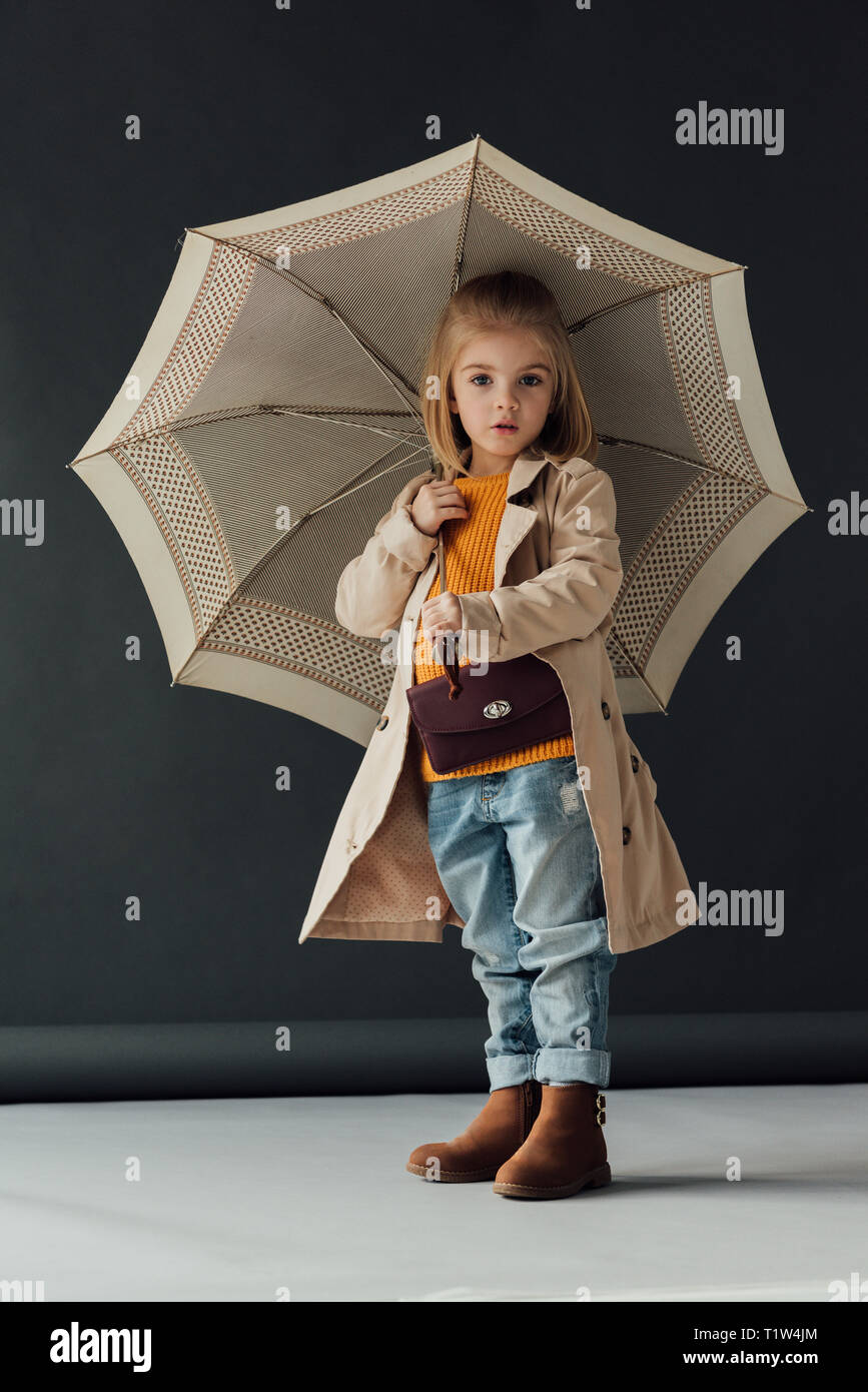 surprised child in trench coat and jeans holding umbrella and looking at camera Stock Photo