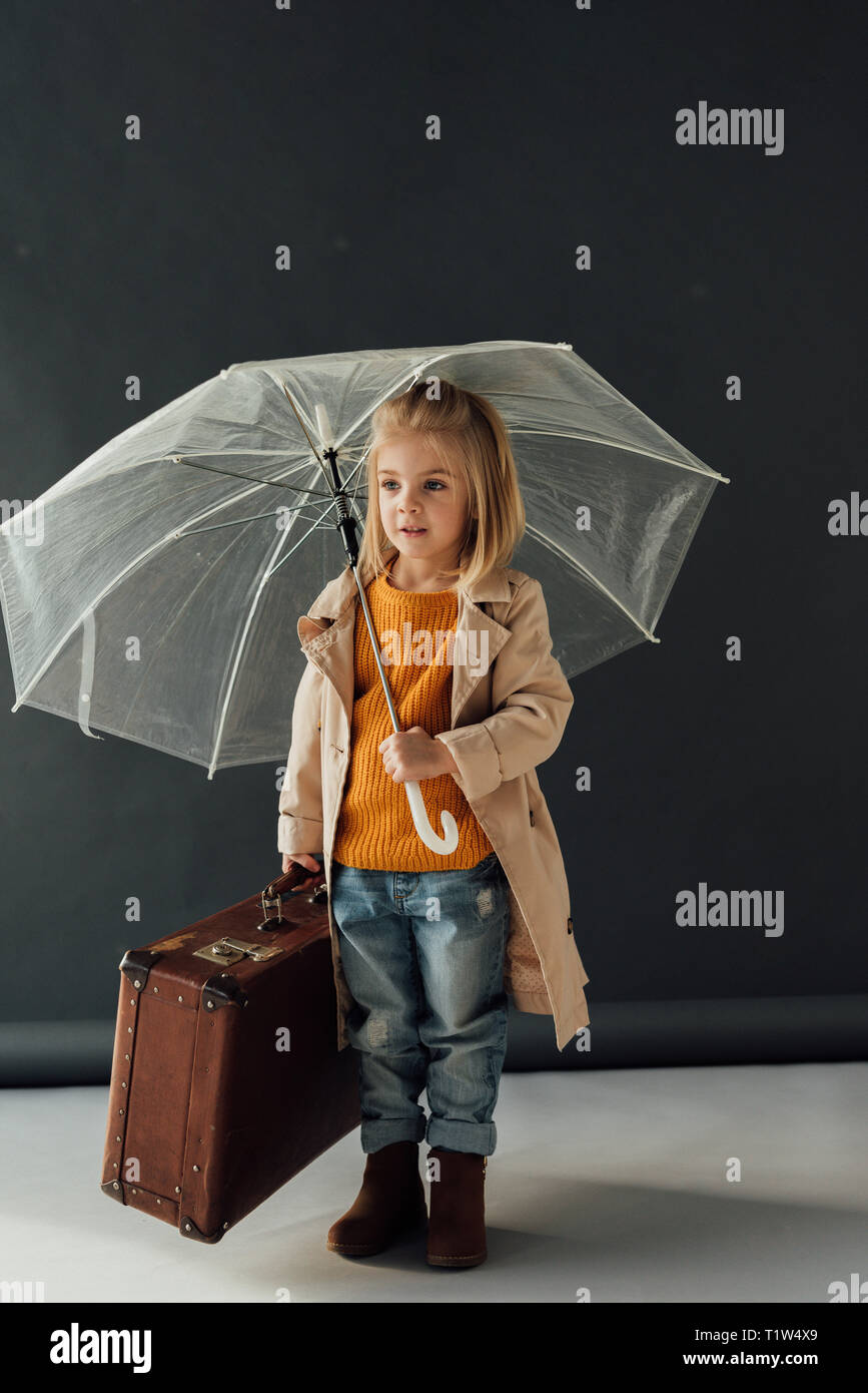 child in trench coat and jeans holding umbrella and leather suitcase on black background Stock Photo