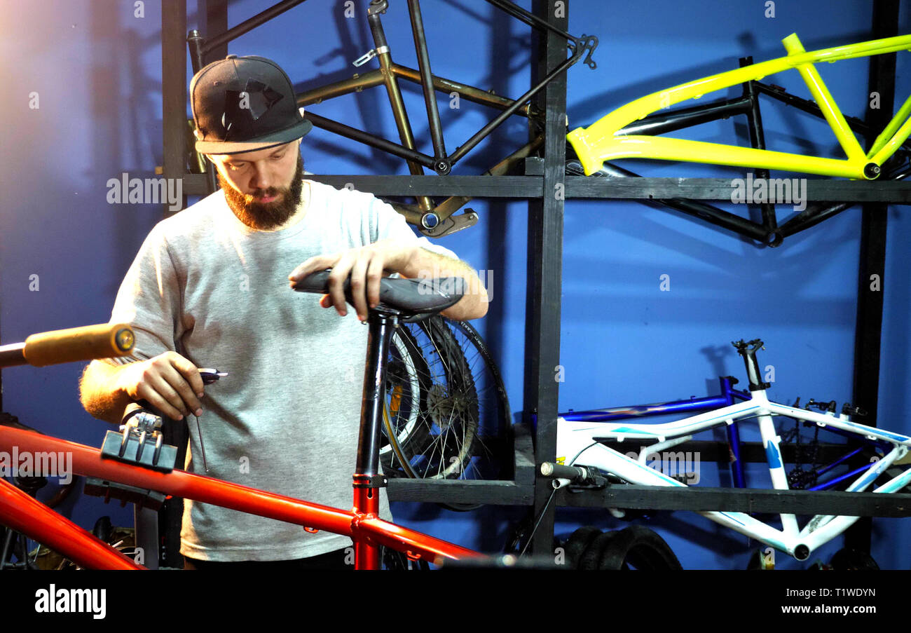 A man bike mechanic assembles a mountain bike in his workshop. A man wearing a cap with a beard. The mechanic fastens the bicycle seat to the bicycle  Stock Photo