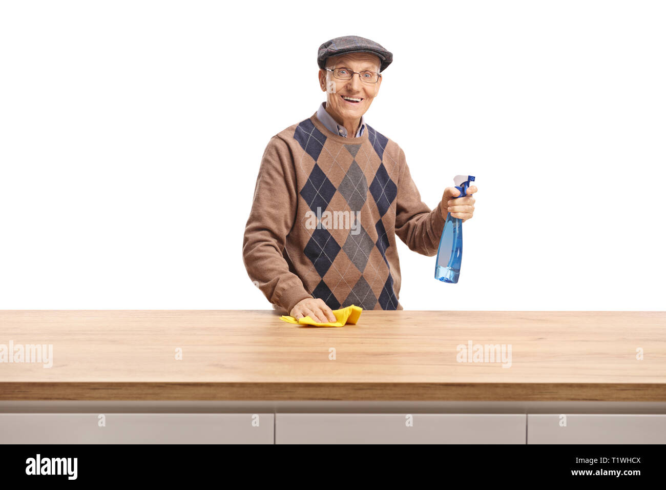 Elderly man with a cloth and cleaning spray standing behind a wooden counter and smiling at the camera isolated on white background Stock Photo