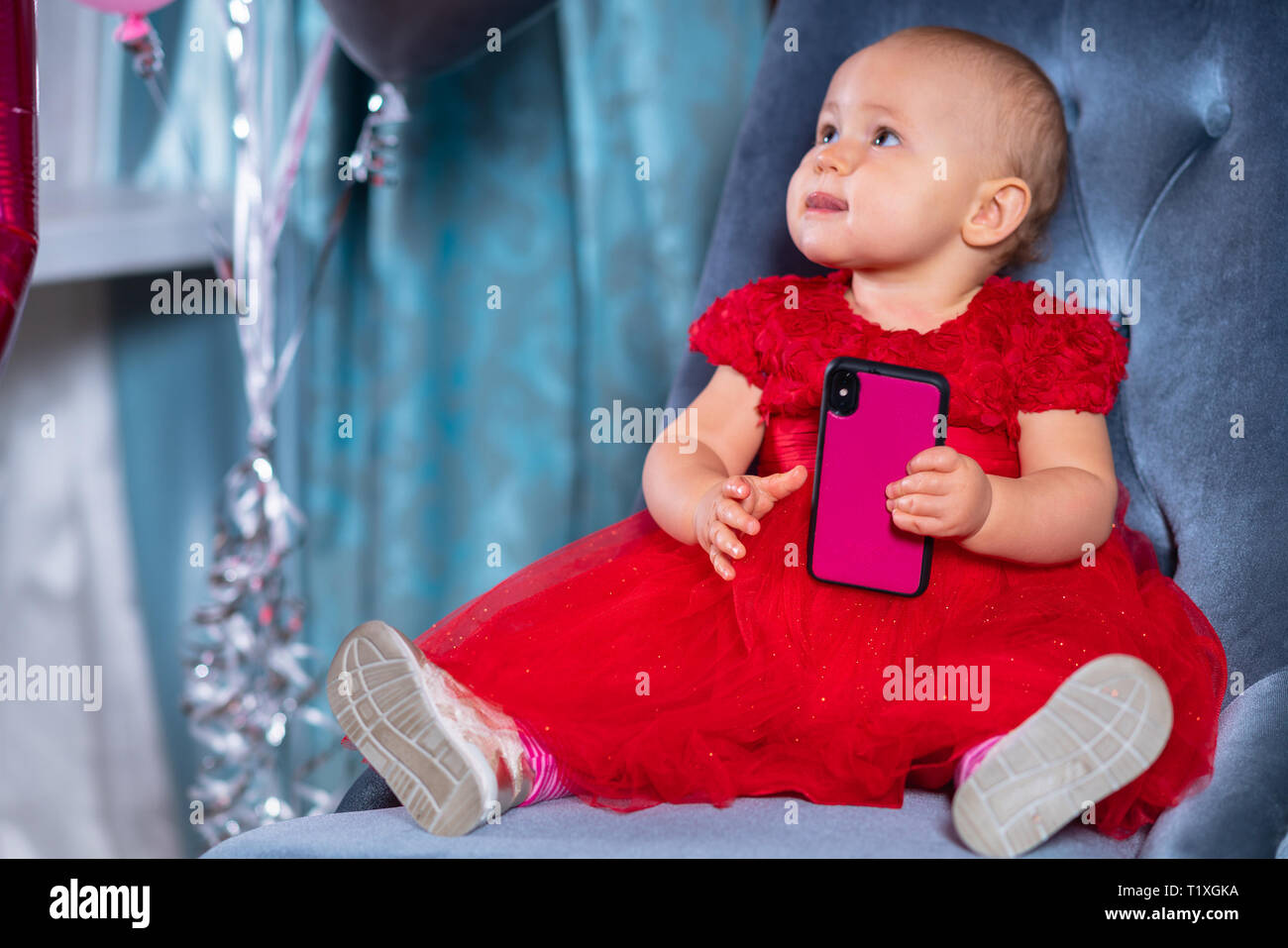 Cute little baby girl in festive holiday red dress, sitting on blue velvet chair and playing with smartphone in her hands Stock Photo