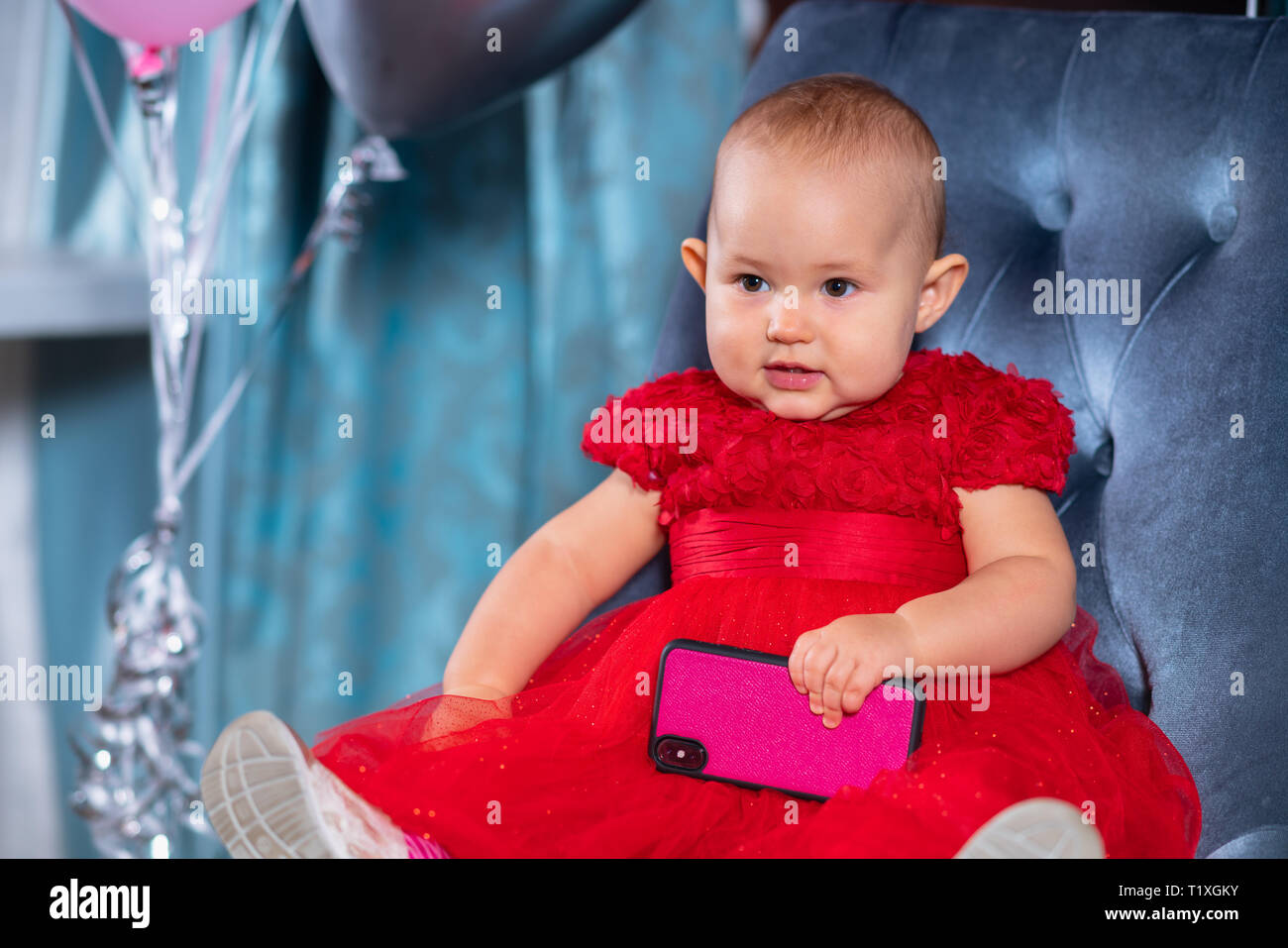Cute little child girl in festive red dress with big smartphone in her hand, sitting on blue velvet chair with balloons in background and smiling Stock Photo