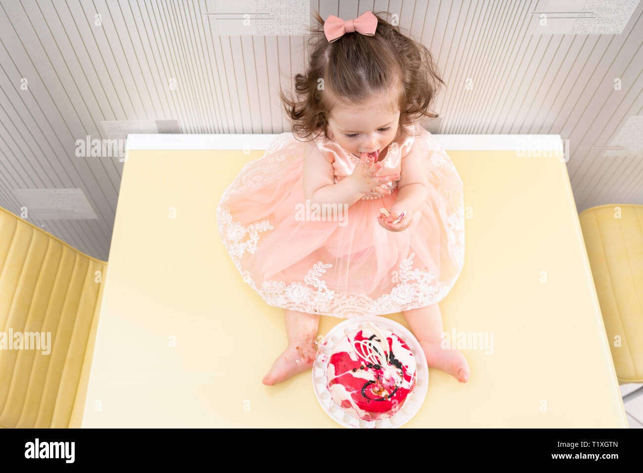 Little baby girl in pink dress sitting in front of the cake on yellow ...