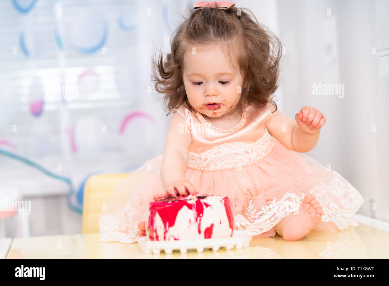 Messy pretty little girl in a pink frock eating a birthday cake with jam smeared around her mouth and sticky hands as she sits along it on a table Stock Photo