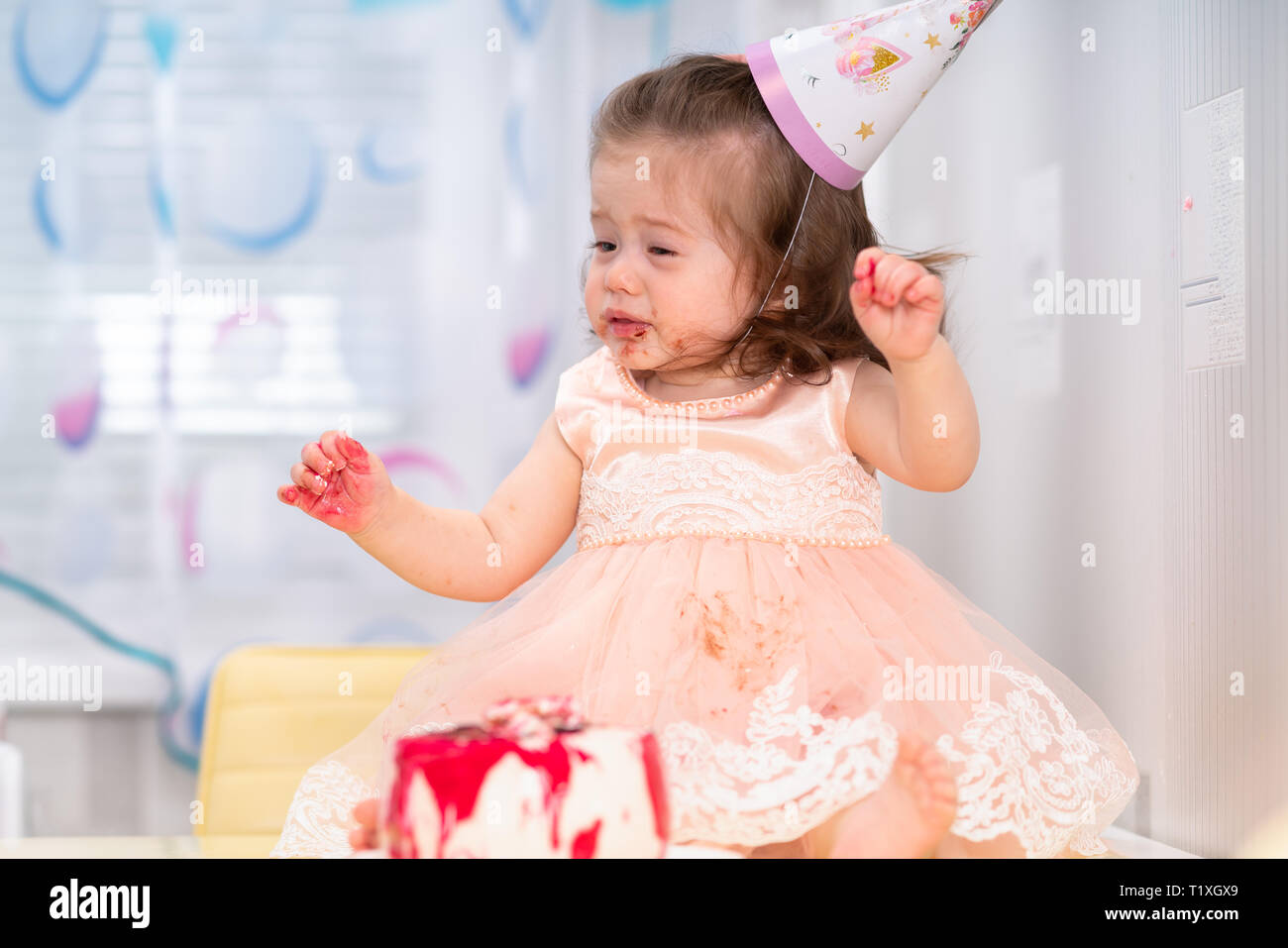 Little girl enjoying her birthday party sitting in front of the cake with sticky hands and face wearing a hat and pink dress Stock Photo