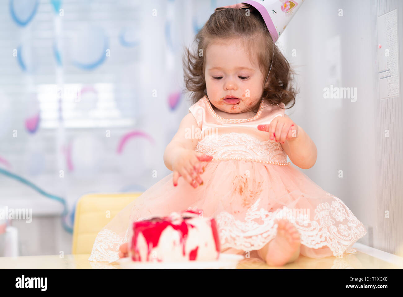 Cute sticky messy little girl in a pink dress eating her birthday cake wearing a party hat Stock Photo