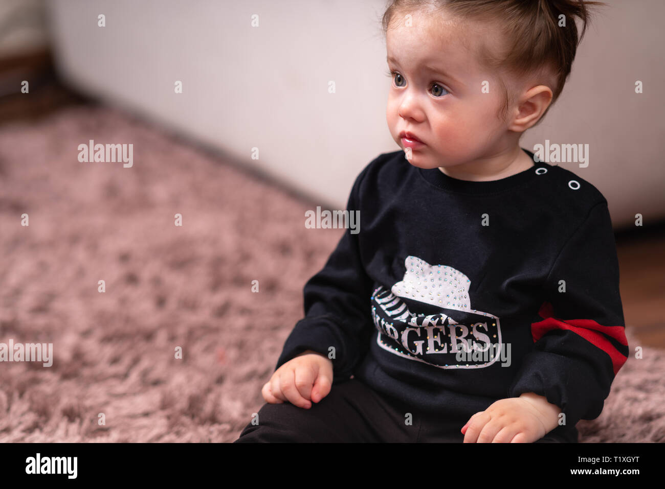 Cute little baby girl in black shirt, sitting on a carpet on floor in living room and looking away. Close-up high angle portrait with copy space Stock Photo