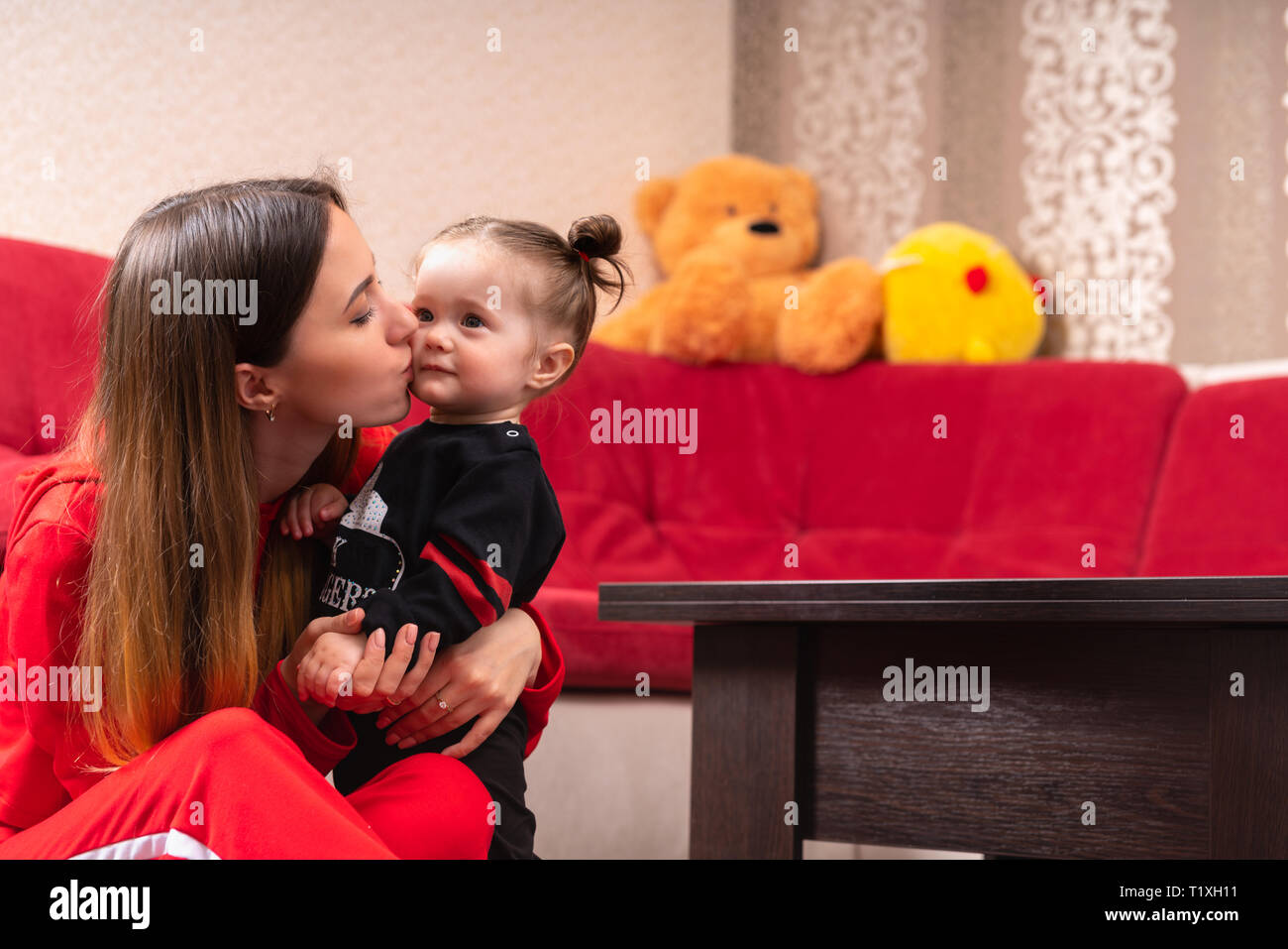 loving-young-mother-kissing-her-baby-daughter-as-she-hugs-her-close-while-sitting-on-the-nursery