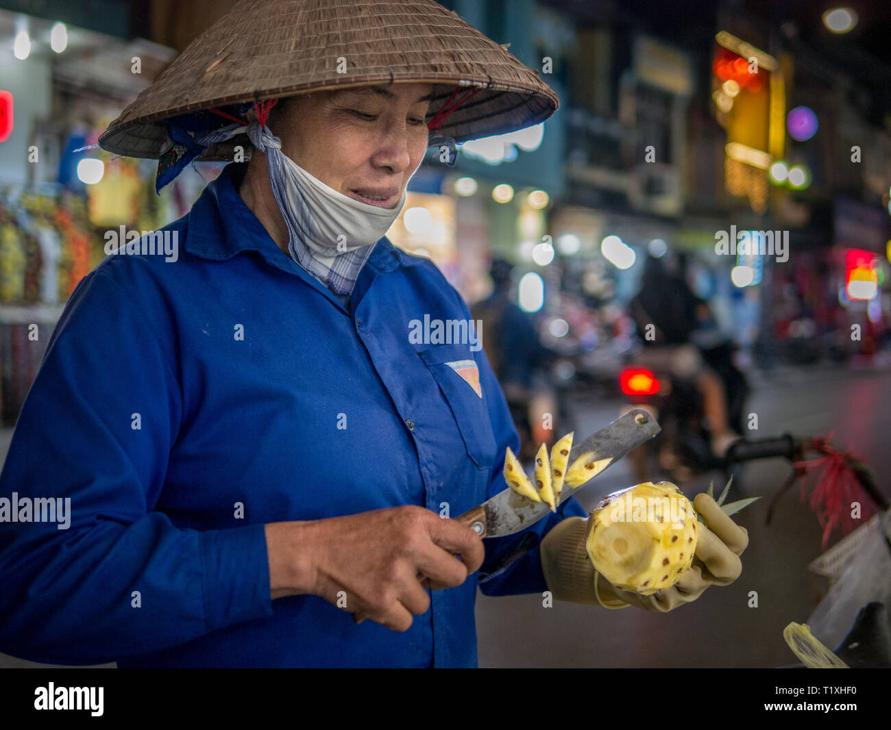 Vietnamese woman with classic conical hat preparing and slicing pineapple at night market in Hanoi, Vietnam Stock Photo
