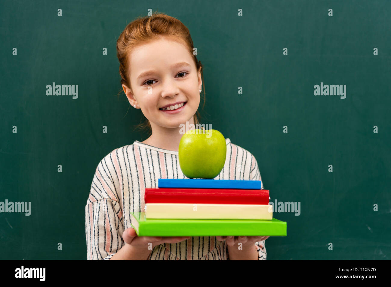 Cheerful ginger shoolgirl holding books and green apple in front of ...